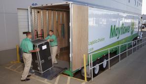 Two men in green shirts move a large case out of a Mayflower moving van.